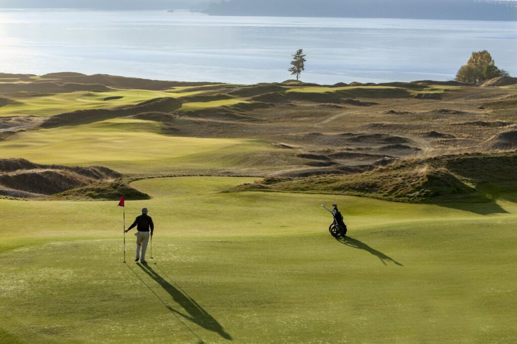 tacoma-chambers-bay-golf-1 - junglecity.com Golfer near a red-flagged hole on a sunny coastal golf course; a golf bag trolley sits on the rough nearby.