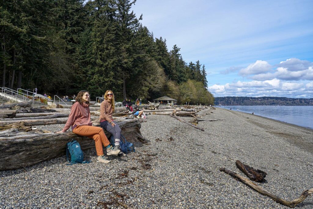 tacoma-point-defiance-park-4 - junglecity.com Two people sit on a large driftwood log on a pebbled beach, with forested steps nearby and calm water in the background.