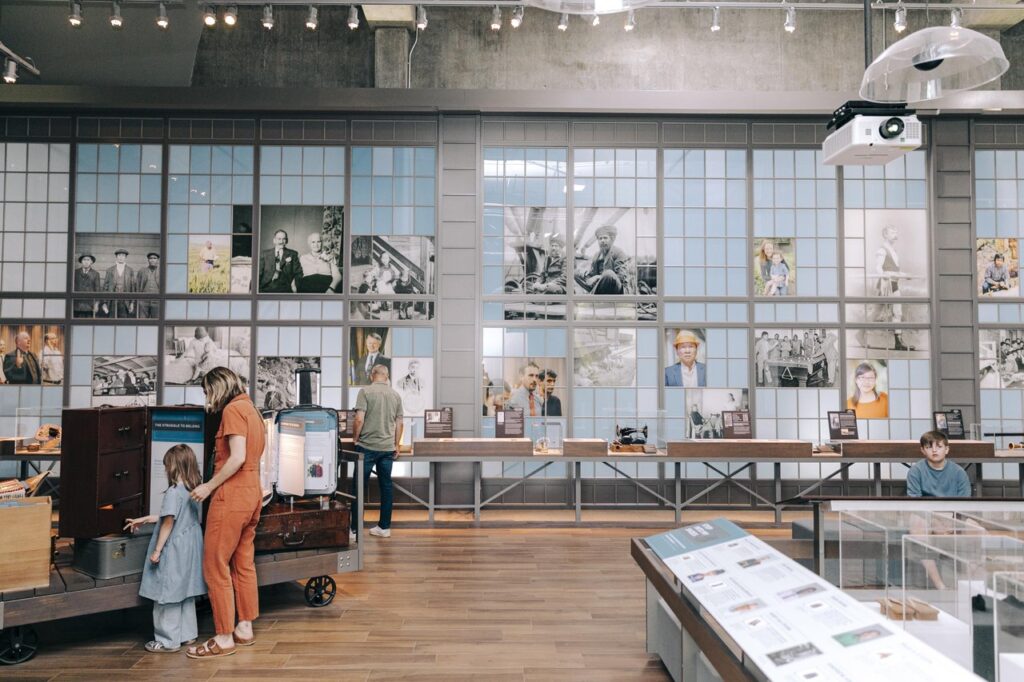 wa-state-history-museum-1 - junglecity.com Museum gallery with a grid wall of photos, a mother in orange helping a child near a vintage luggage cart on the left, and a boy seated at right.