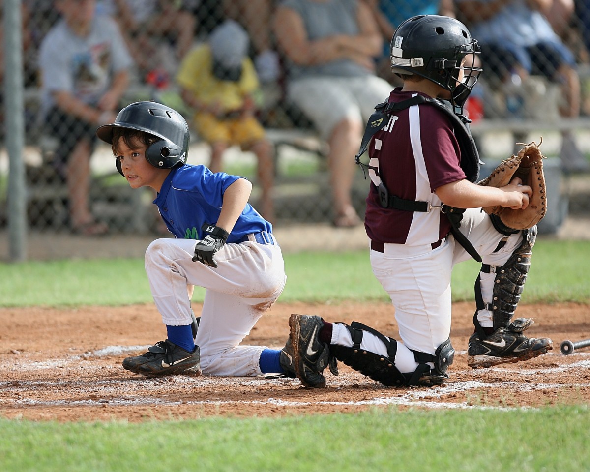 Two young baseball players on a dirt field: a blue-jersey runner slides into base while a catcher in maroon stands nearby with a glove, spectators blurred in the background.