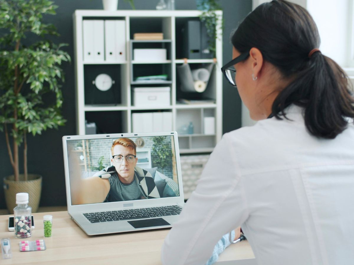 Clinician in a white coat talks to a patient on a laptop during a virtual consult, meds and supplies on the desk.