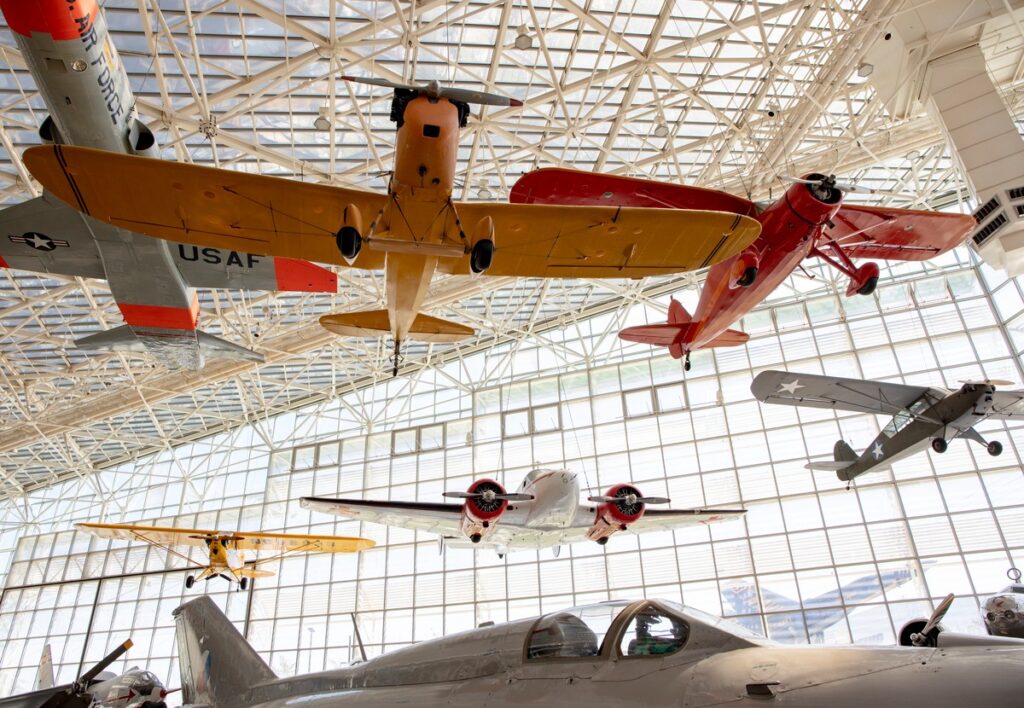 museum-of-flight-south-wall-1 - junglecity.com Hangar ceiling with several vintage propeller airplanes suspended above a gray fighter jet in the foreground in a museum setting