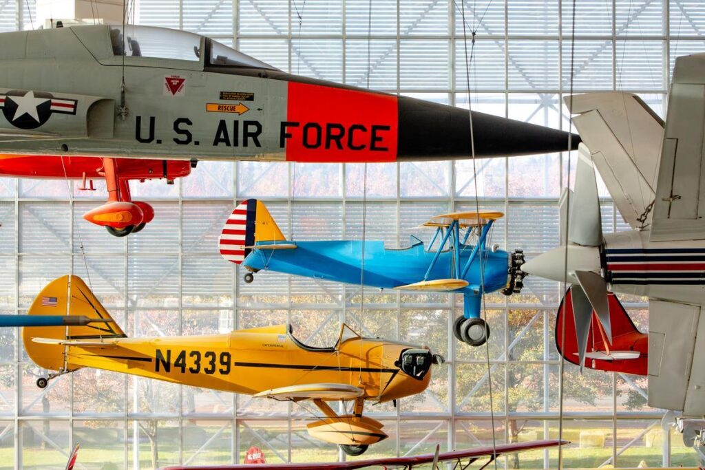 Vintage aircraft display in a hangar, with a U.S. Air Force jet overhead and colorful biplanes and a yellow monoplane below.