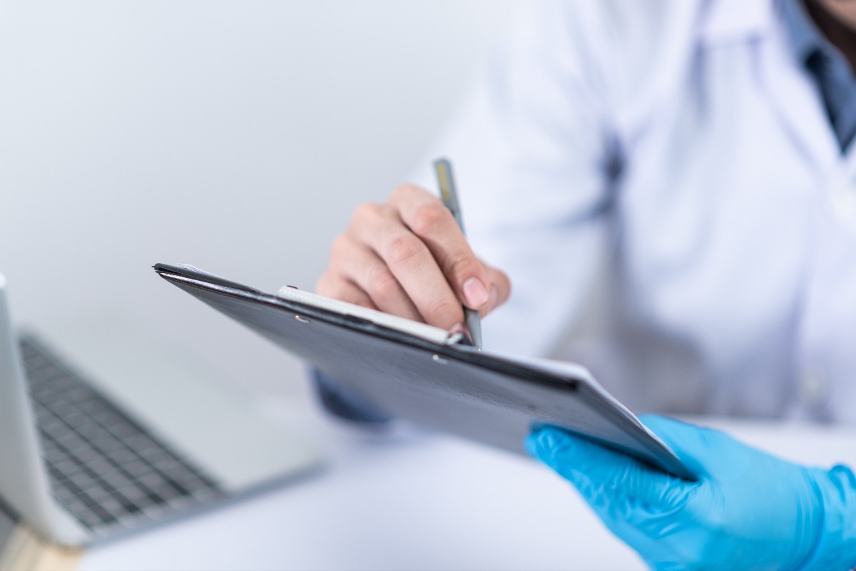 Medical professional in a white coat wearing blue gloves writes on a clipboard with a pencil, laptop visible in the background