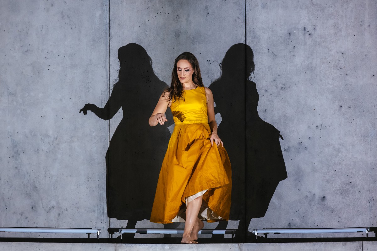 Woman in a bright yellow sleeveless dress walks barefoot on a stage, casting two shadows on a gray concrete wall.