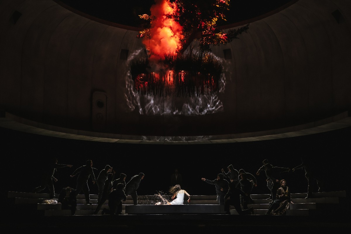 seattle-opera-salome - junglecity.com Dramatic stage scene with a dancer in white center, surrounded by silhouetted performers, as a red flame-like projection erupts overhead.