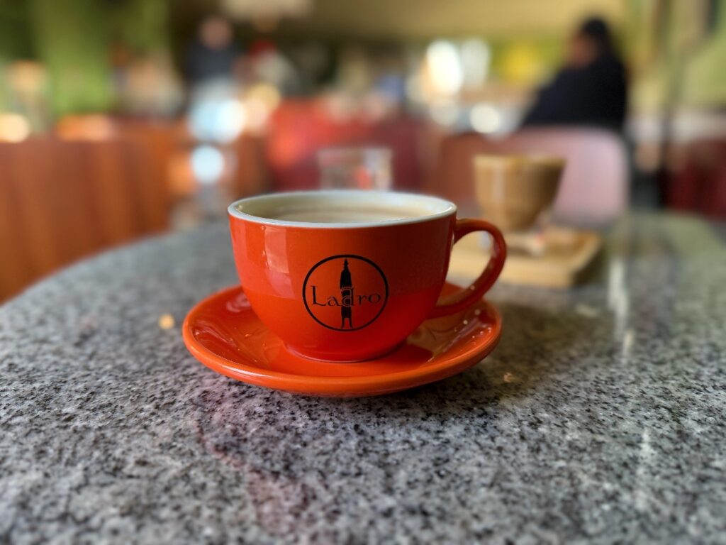 Red ceramic coffee cup on a matching saucer sits on a speckled tabletop in a cafe; cup features a black Ladro logo.
