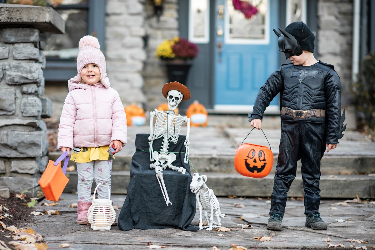 Two kids in costume stand on a porch: a girl in a pink winter coat and hat with an orange pumpkin bucket, a boy dressed as Batman with a pumpkin bucket, beside a skeleton decoration on a black-draped chair.