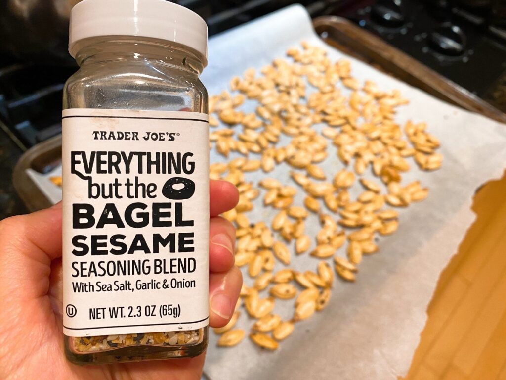 Bottle of Trader Joe's Everything but the Bagel Sesame Seasoning Blend held in hand with sesame seeds spread on parchment in background. Includes label text.
