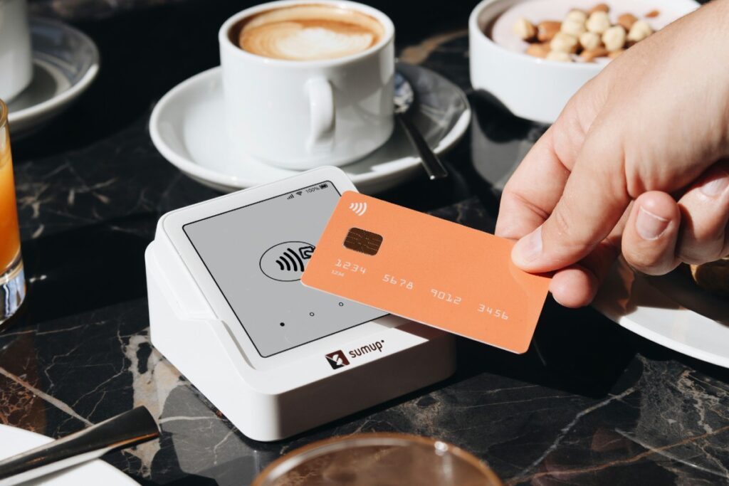 Hand tapping an orange contactless card on a white SumUp payment terminal at a cafe table, with coffee cup nearby.