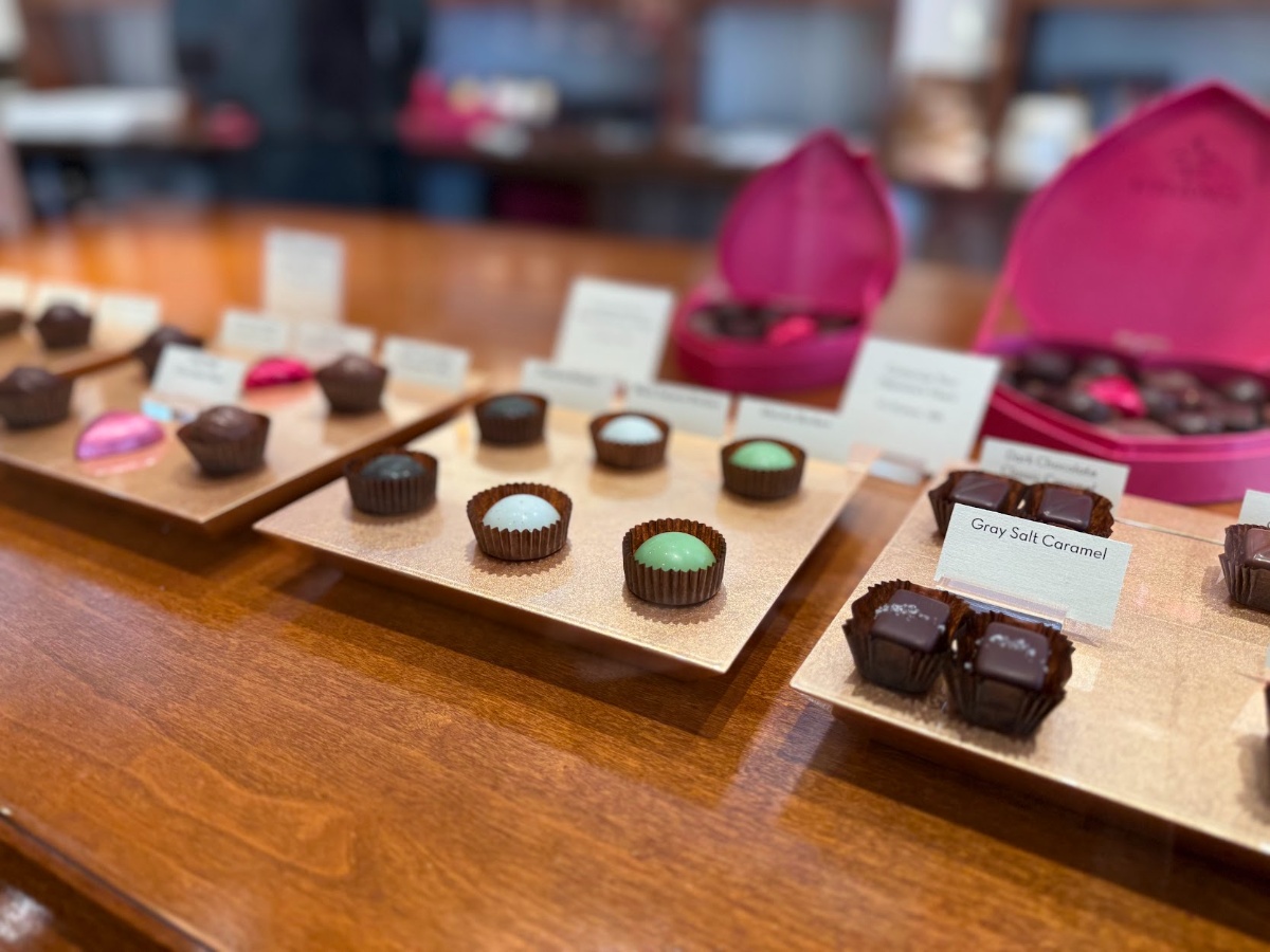 Assorted chocolate truffles on gold display trays with flavor labels at a shop counter.
