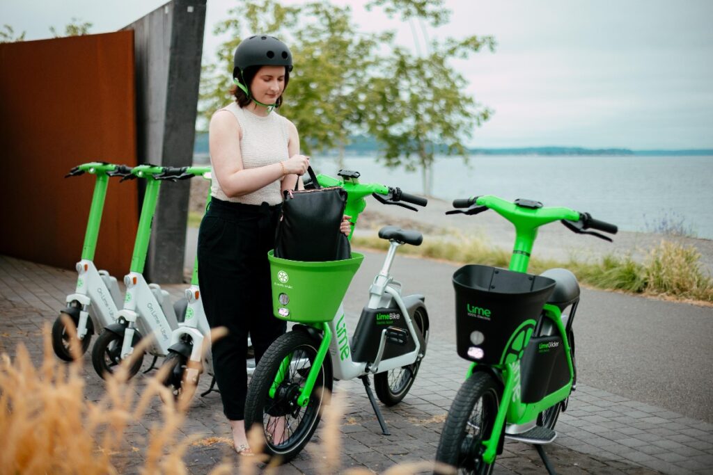 A woman wearing a helmet stands by bright green Lime bikes parked along a lakeside path, holding a black bag.
