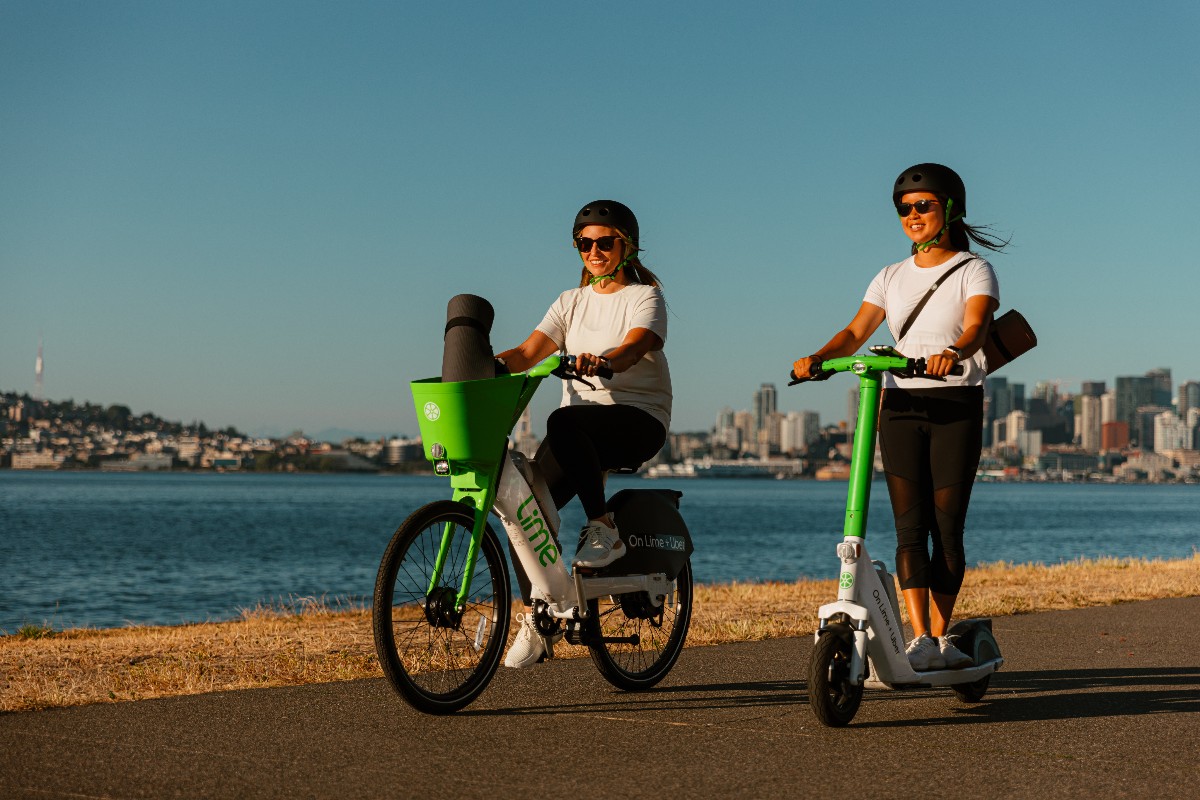 Two women wearing helmets ride lime-green e-bikes along a waterfront with a city skyline in the background.