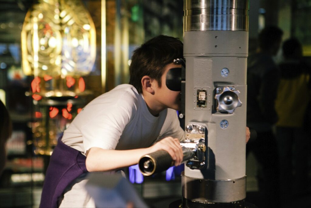 Person peering into a large gray telescope mounted on a metal pedestal in a museum-like setting.
