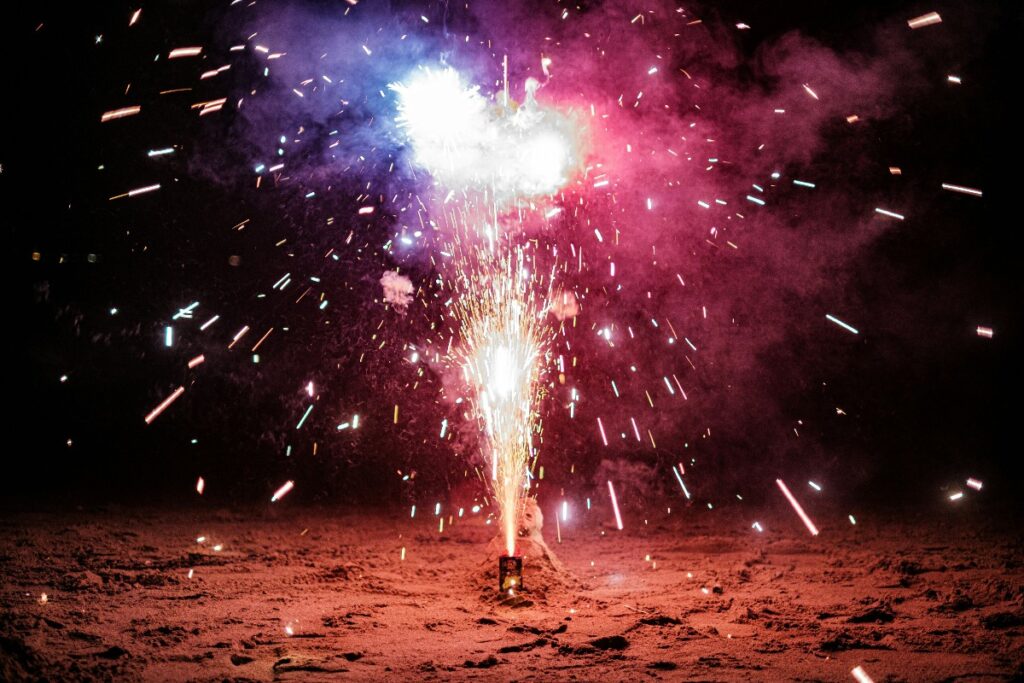 Firework fountain blasting bright sparks on the ground at night with colored smoke in the background.