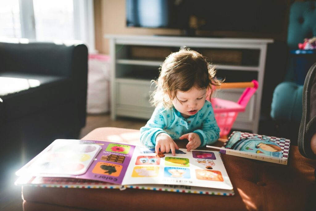 Toddler in blue pajamas sits at a wooden table, pointing at a bright, colorful picture book laid open before them.