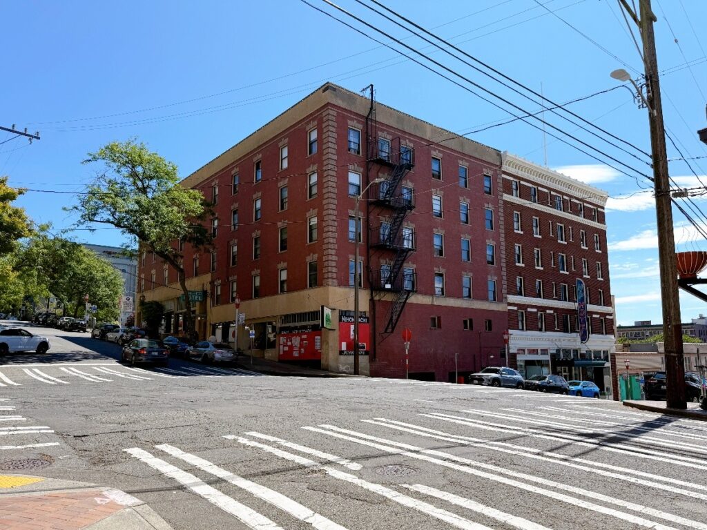 Red brick, four-story urban building at a street corner with a black fire escape on its side, ground-level storefronts, and a crosswalk in the foreground under a clear blue sky.