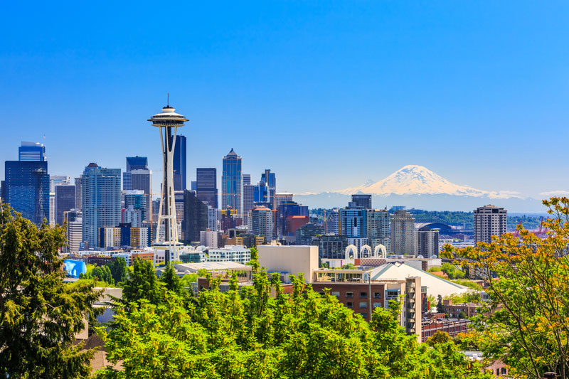 Seattle skyline with the Space Needle and distant Mt. Rainier under a clear blue sky, seen from a green foreground park area.