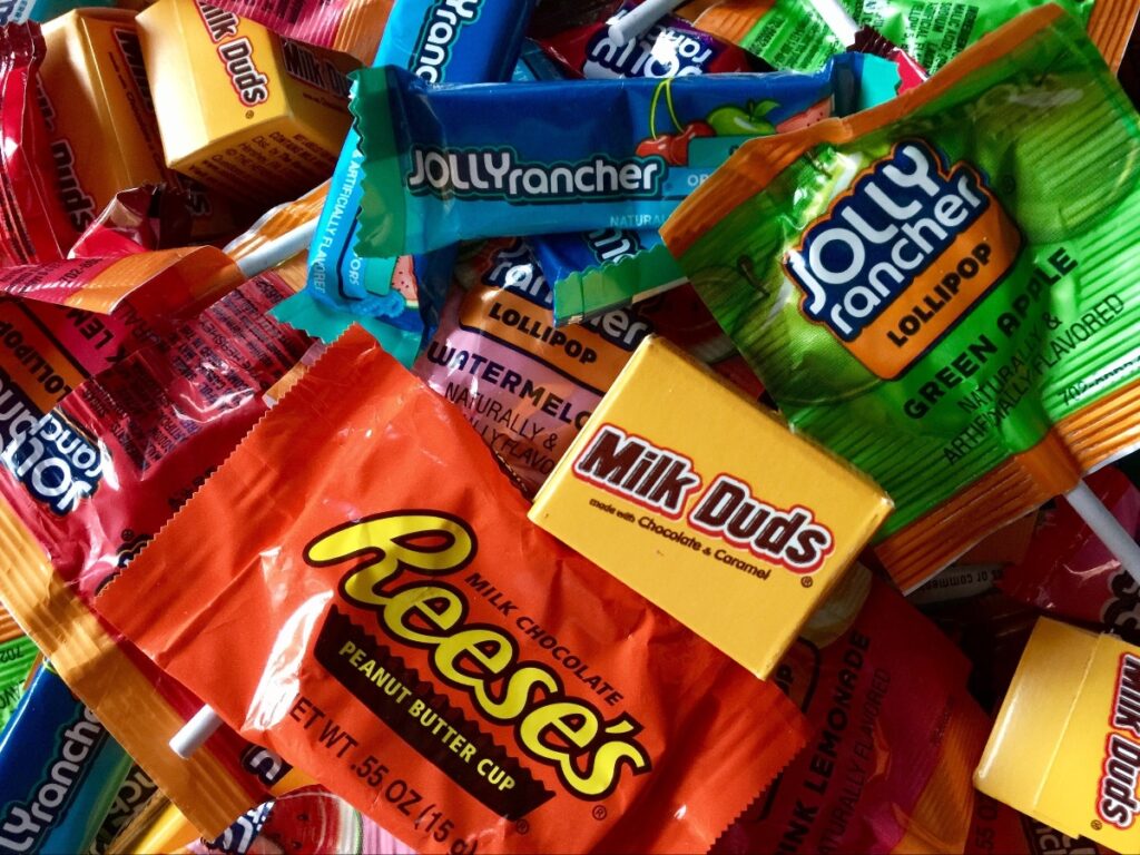Pile of assorted candy wrappers: Reese's, Milk Duds, Jolly Rancher (green apple), and other colorful treats mixed together