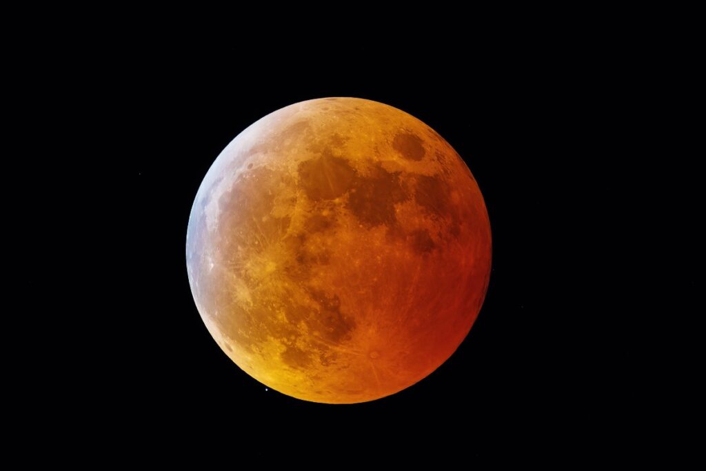 Coppery total lunar eclipse: the Moon glowing orange-red against a dark night sky, with visible craters and shading across its surface.