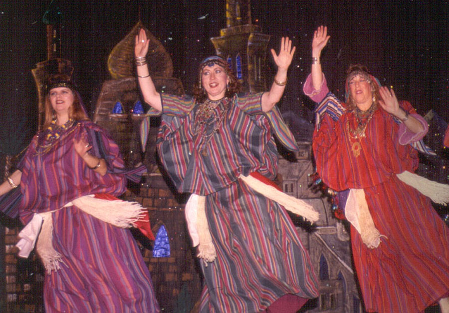 arab - junglecity.com Three women in colorful striped robes dance on a stage with a castle backdrop, hands raised above their heads.