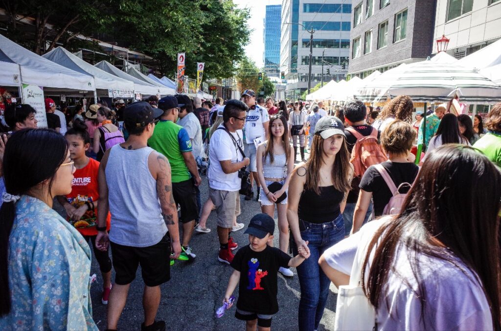 Crowded urban street festival with white vendor tents, people browsing stalls, and tall buildings in the background.