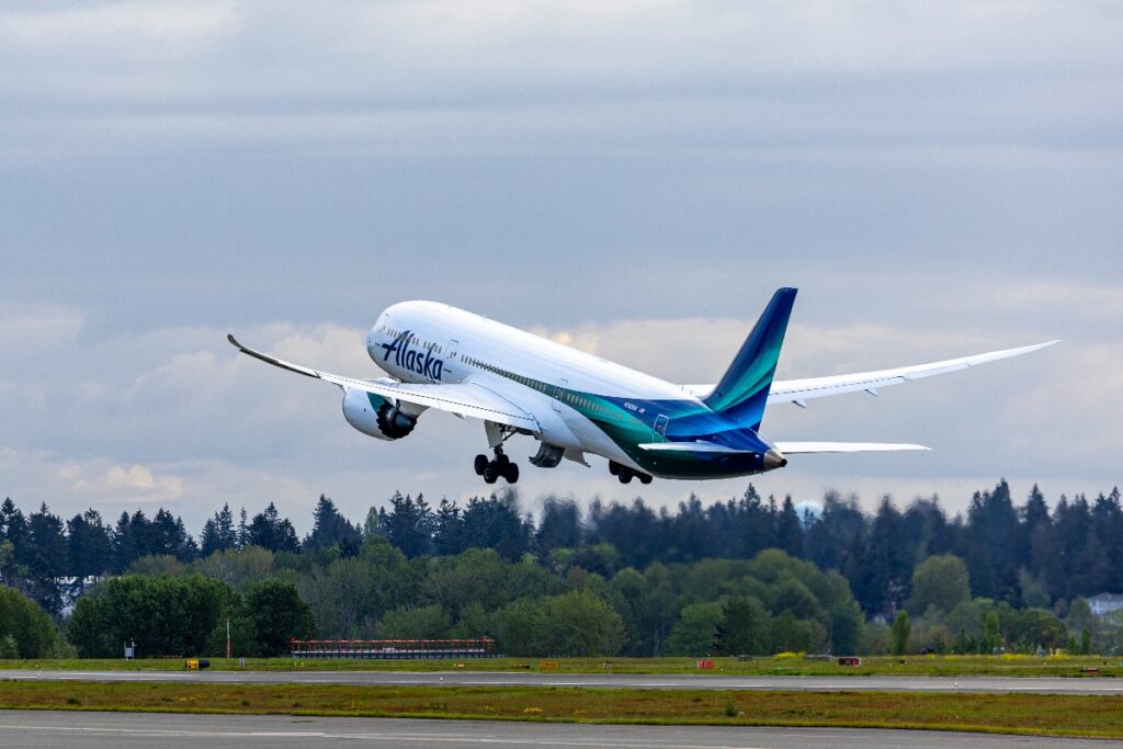 Alaska Airlines jet lifting off from a runway with landing gear retracting, blue-teal tail visible against a cloudy sky and trees in the background.