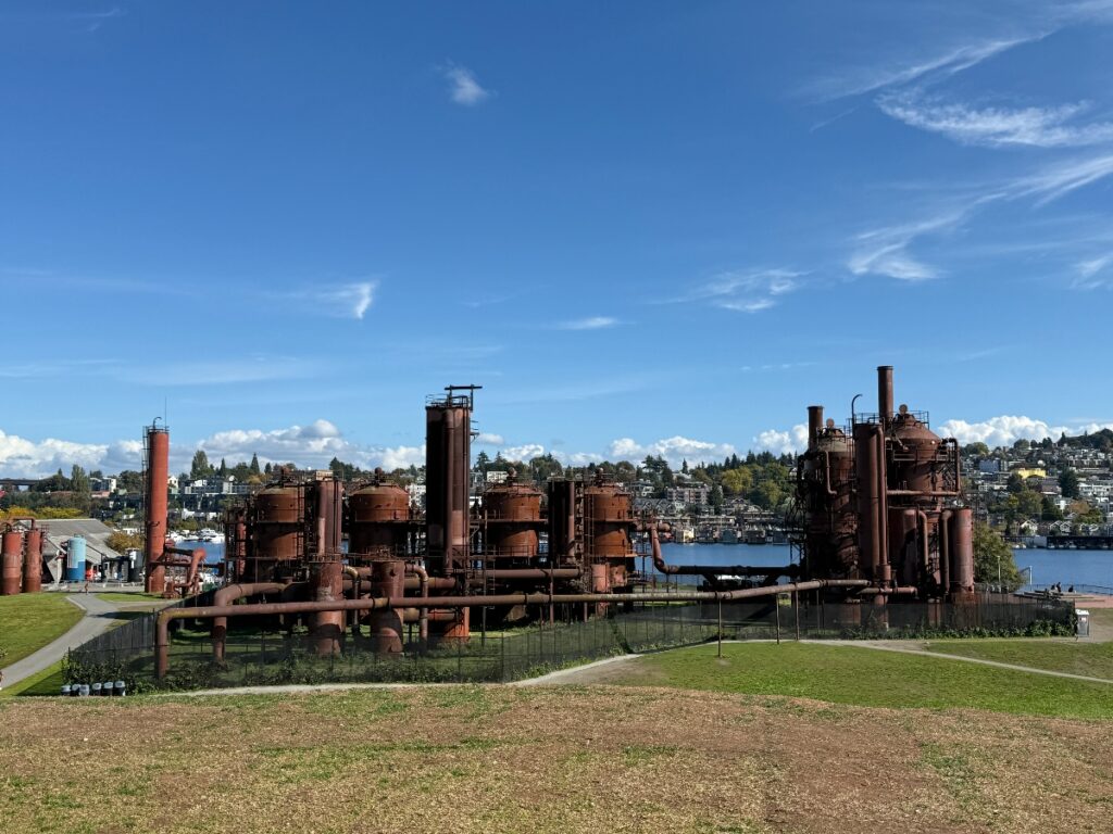 Rusted industrial plant by a lake with interconnected pipes and cylindrical towers under a blue sky.