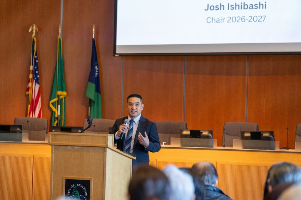 Male speaker in a suit at a wooden podium addressing an audience in a conference room, with flags in the background and a presentation slide reading 'Josh Ishibashi, Chair 2026-2027'.