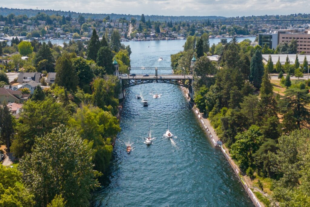 Aerial view of a canal with several boats, crossing under a metal bridge flanked by trees and a city on both banks.
