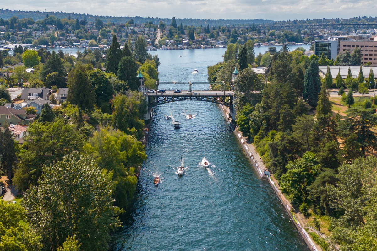 Aerial view of a canal with several boats, crossing under a metal bridge flanked by trees and a city on both banks.