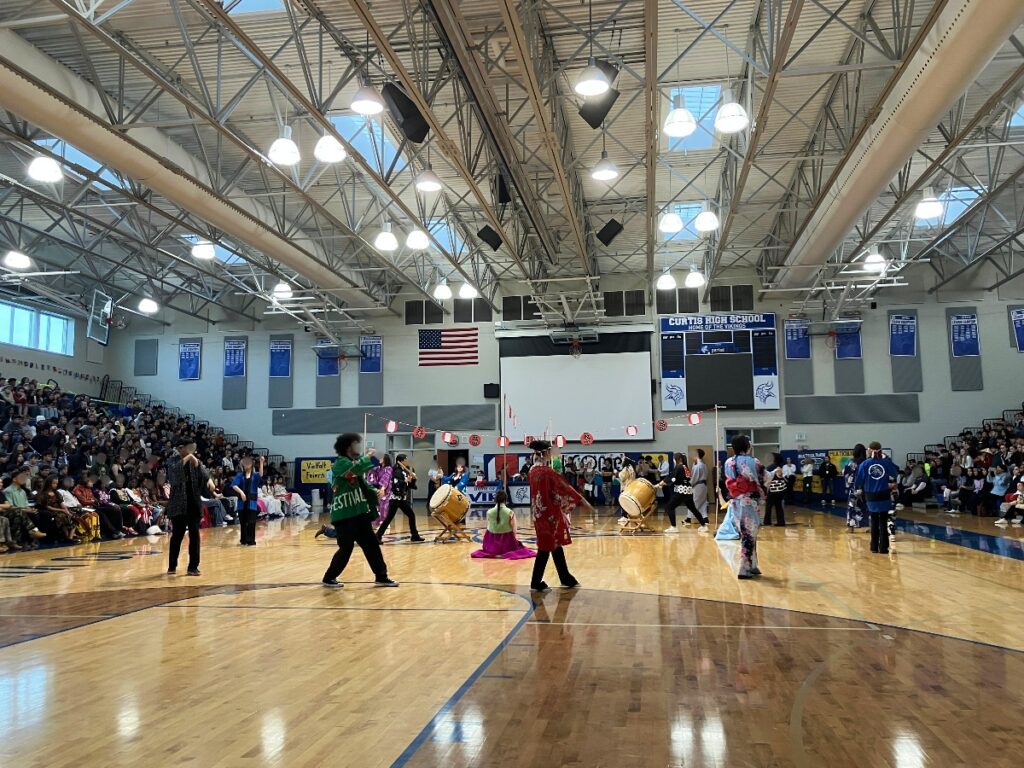 Crowd sits in bleachers as performers in colorful costumes play drums in a school gymnasium.”] ,