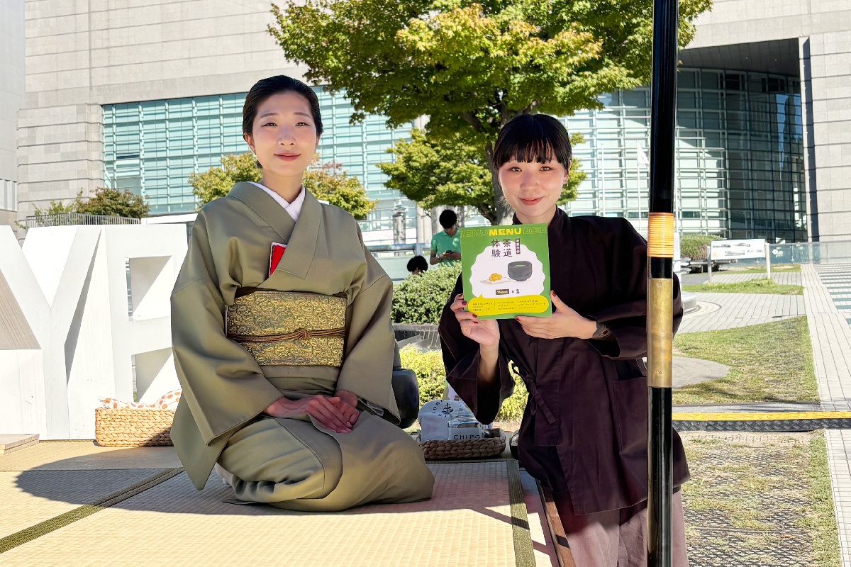 Two women in kimonos sit on a tatami mat outdoors, one in olive green with a gold obi, the other in dark brown holding a bright menu flyer.
