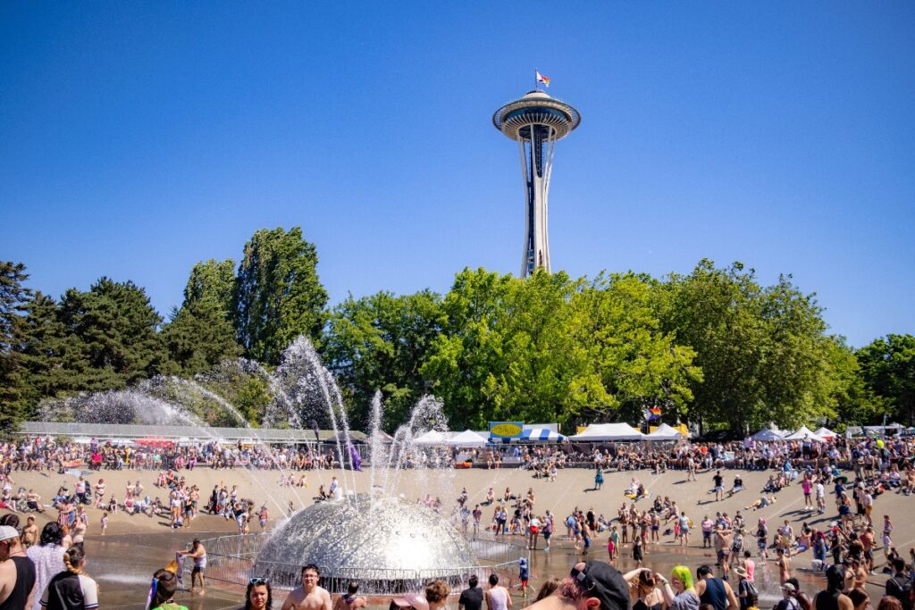 Crowd enjoying a sunny day at a park with a large fountain and the Space Needle in the background.
