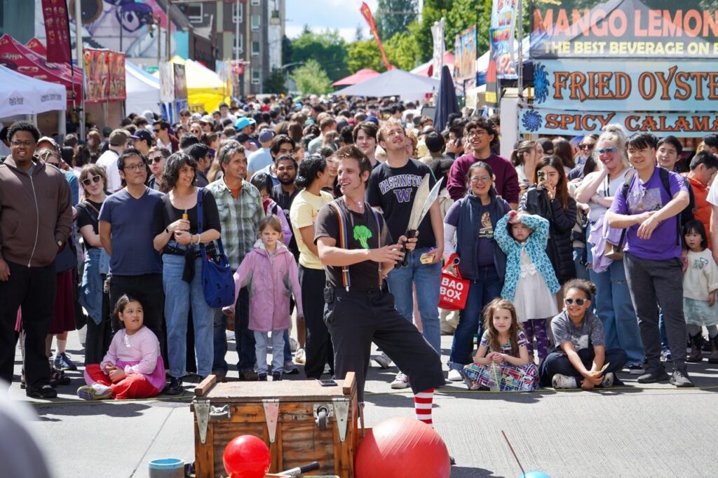Street festival crowd watches a male performer juggling two large knives in front of food stalls and tents.
