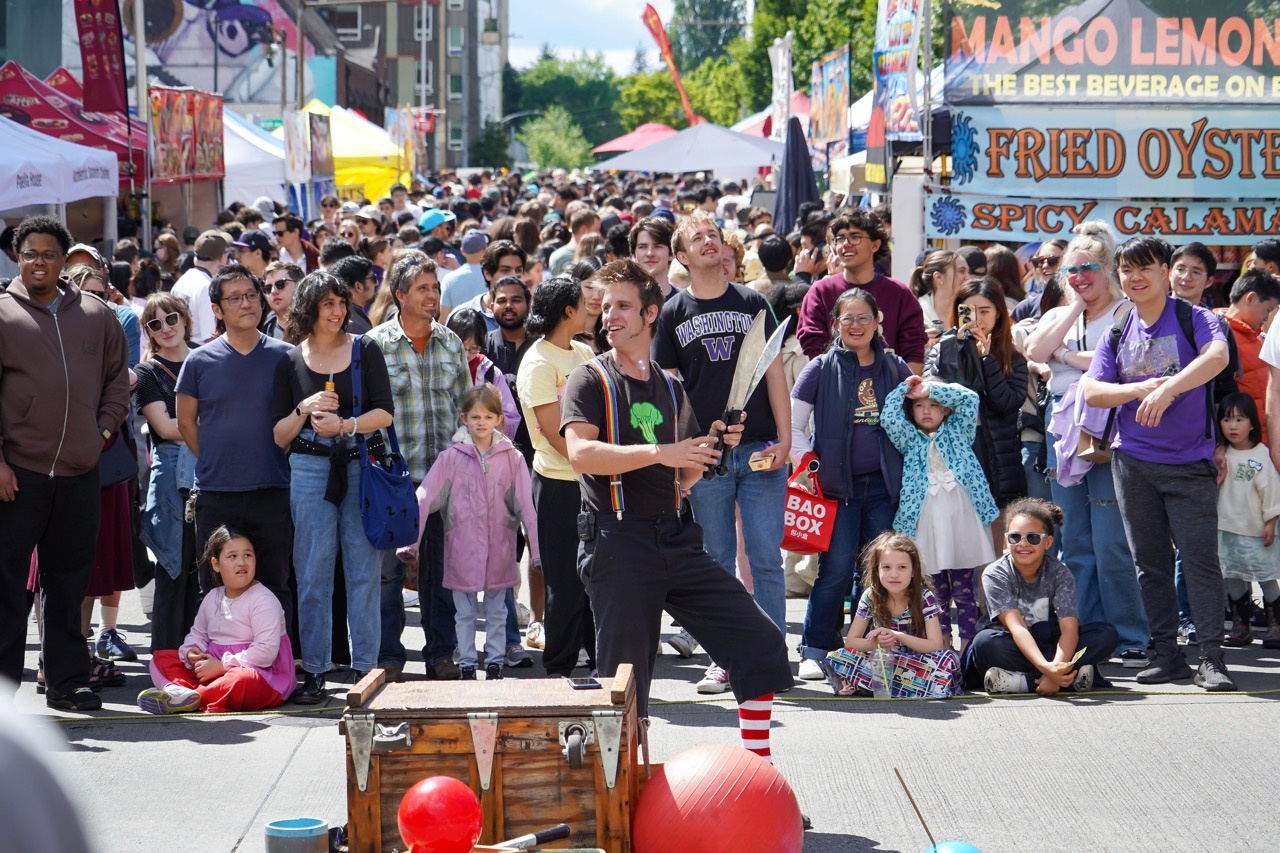 Street festival crowd watches a male performer juggling two large knives in front of food stalls and tents.