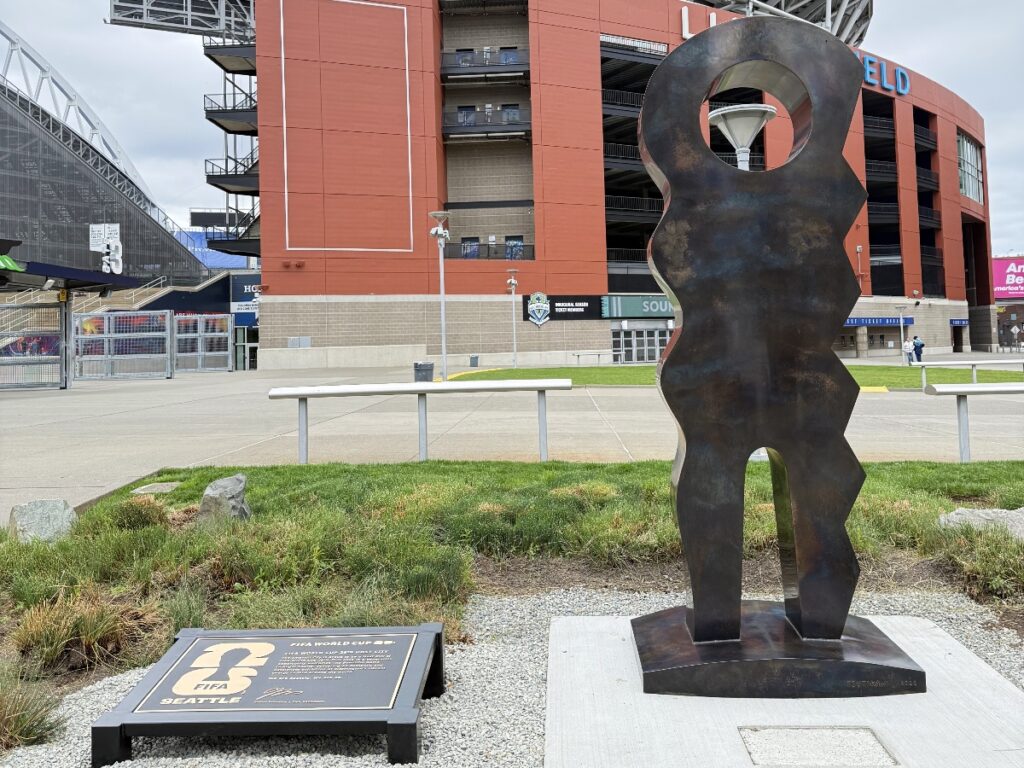 Rusty abstract metal figure sculpture outside a large stadium.