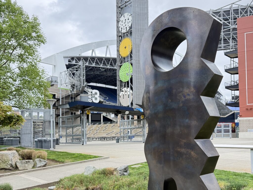 Large rusted metal gear sculpture in foreground with a stadium entrance and tiered seating behind it.