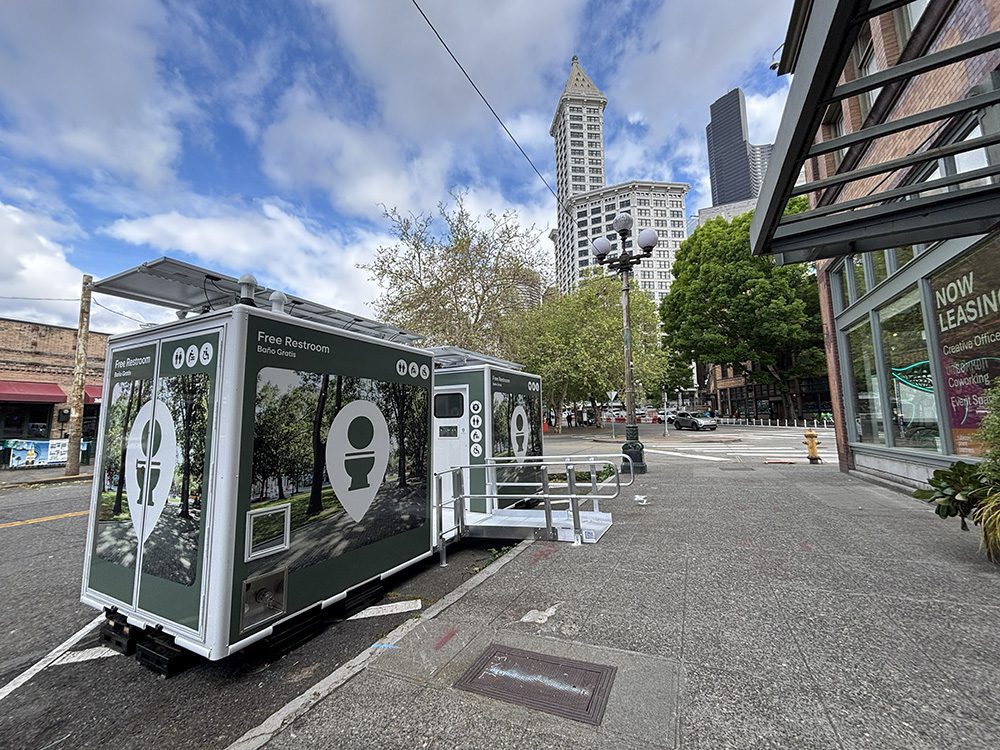 Public portable restroom trailers on a city sidewalk with trees and tall buildings in the background, under a blue sky.