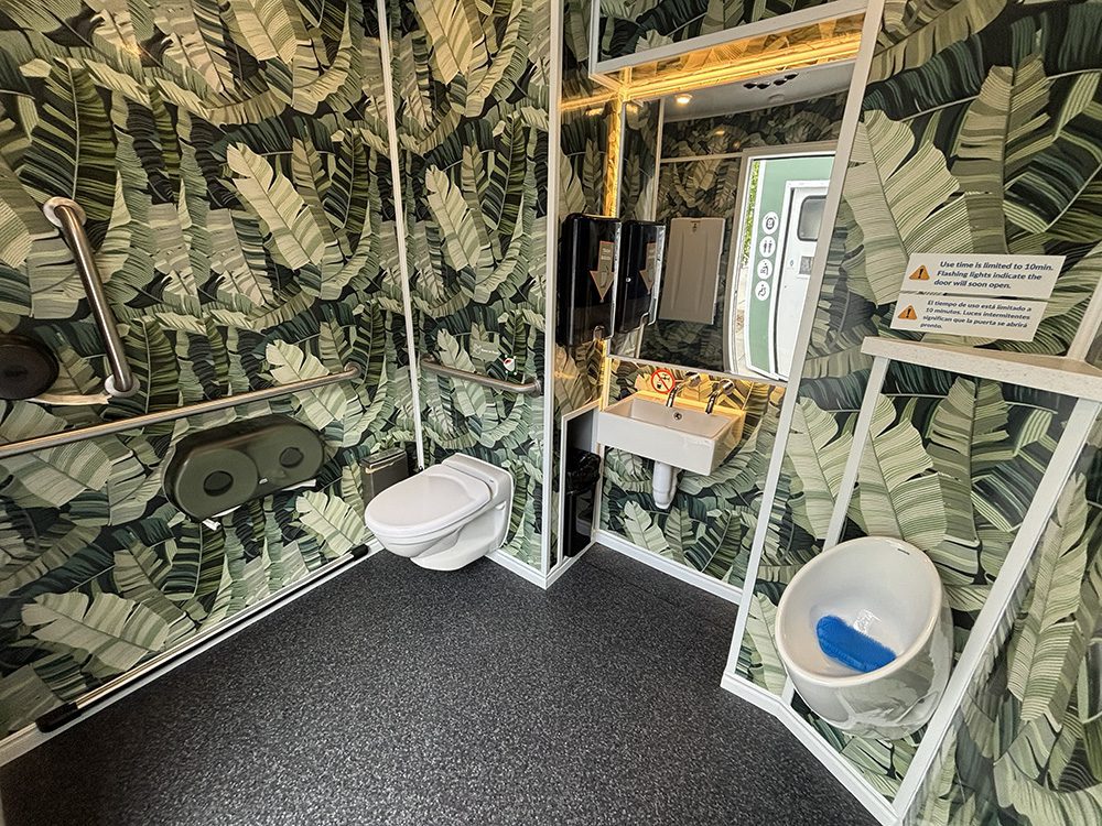 free-restrooms-3 - junglecity.com Public restroom with tropical banana-leaf wallpaper, a wall-mounted toilet, sink, mirror, and grab bars along the walls.