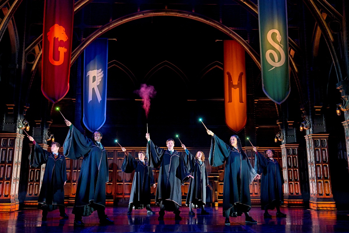 Group of graduates in gowns on stage, each holding a wand with colored smoke, performing a coordinated pose beneath large banners.