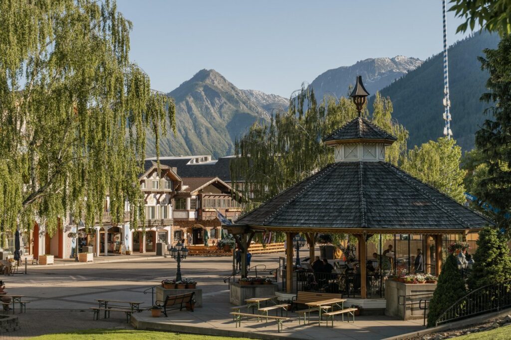 Town square with a wooden pavilion and outdoor seating, set against mountains and alpine buildings under clear skies.