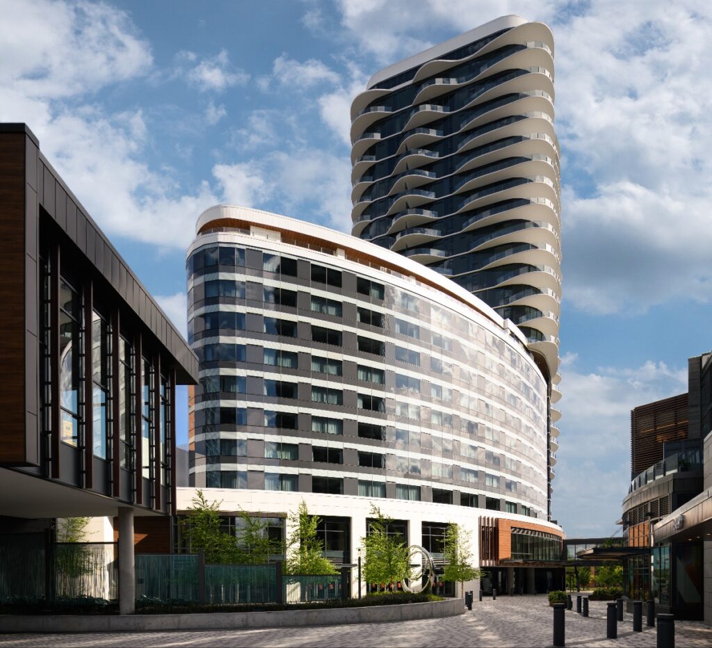 Modern curved glass office building with stacked balconies and a tall rounded tower in a city plaza under a blue sky.