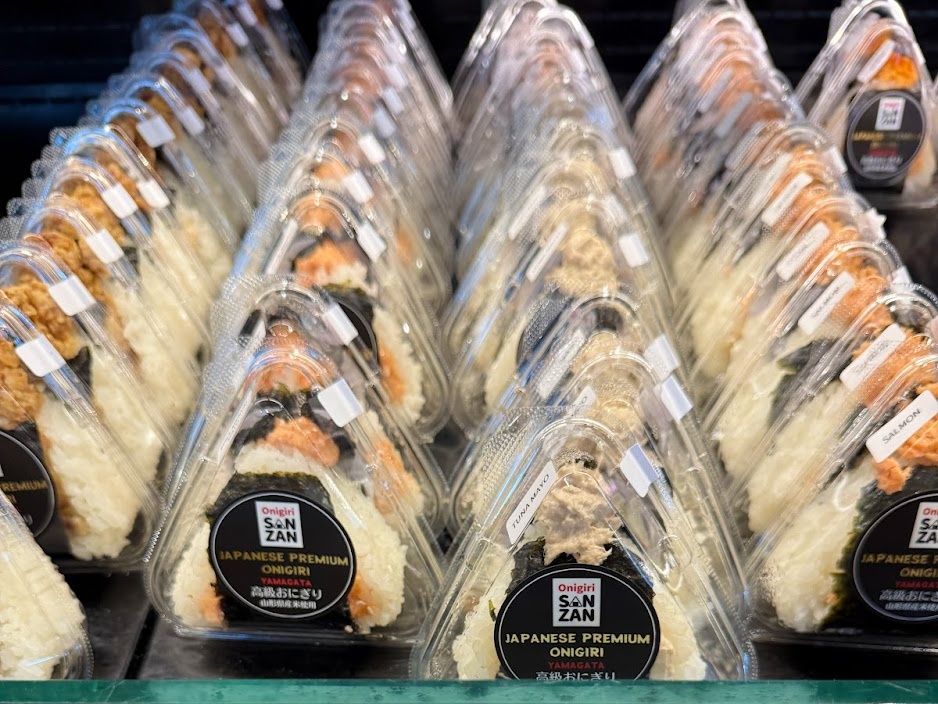 Rows of triangular onigiri rice balls in clear plastic packaging with black labels visible on the front shelves
