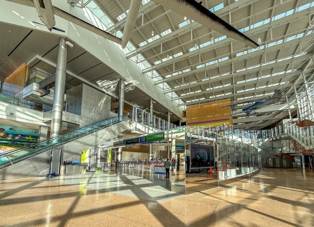 Bright airport terminal with escalators, glass walls, and a hanging model airplane under a skylit roof.