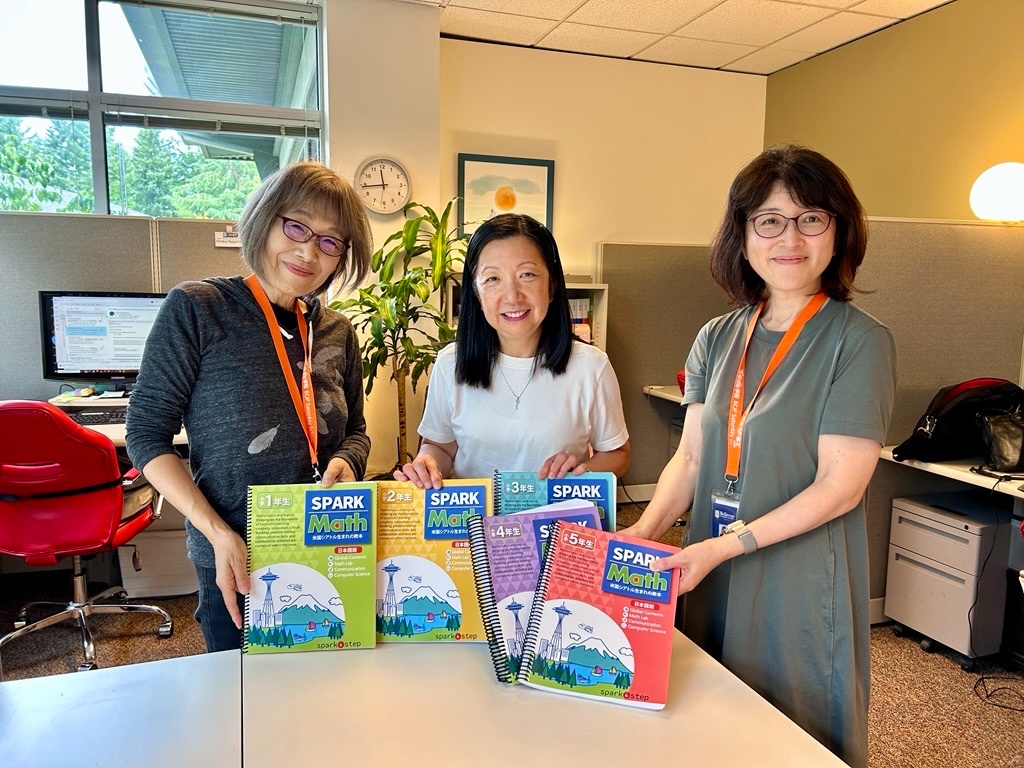 Three women in an office pose behind a table showing colorful Spark Math workbooks.] (optional: includes the visible SPARK Math covers)