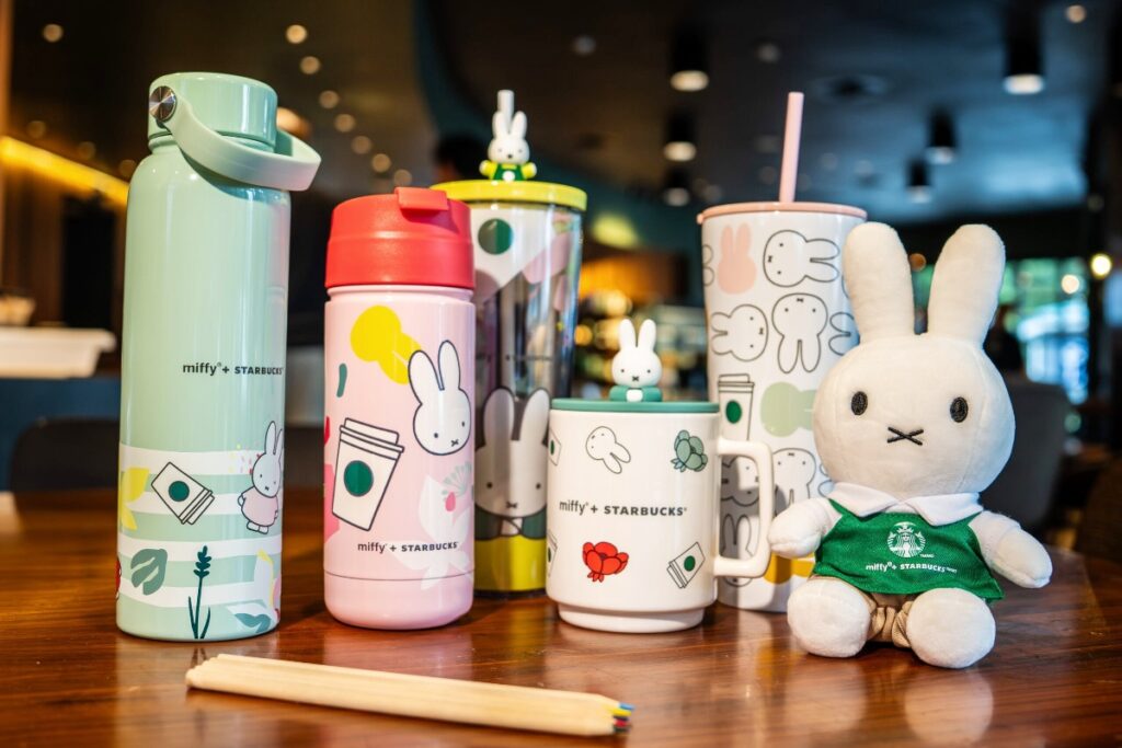 Pastel Miffy x Starbucks bottles and mugs with a white plush bunny in a green apron on a wooden table.