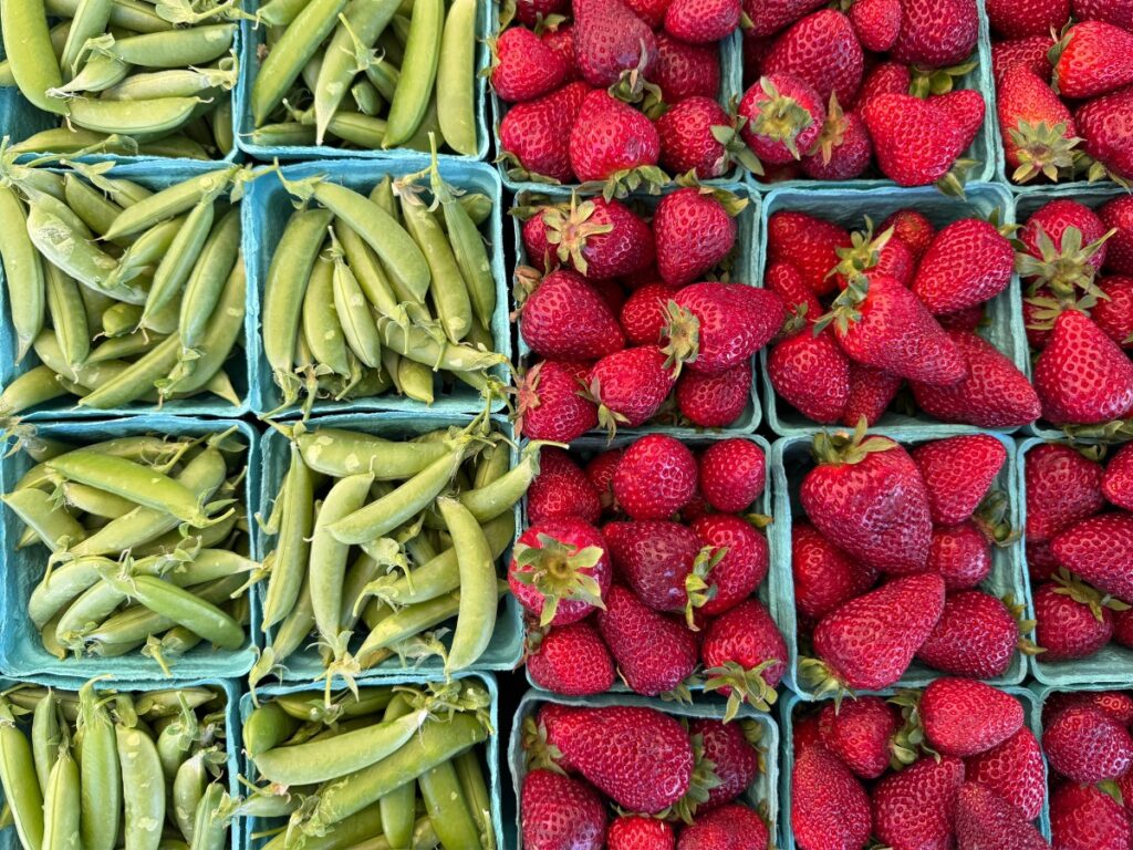 strawberry-1 - junglecity.com Top-down view of fresh green pea pods in blue crates and bright red strawberries in adjacent crates.