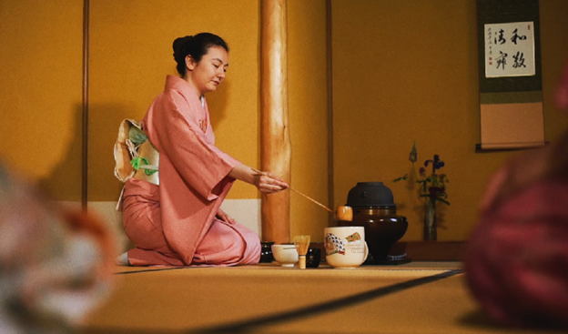 Woman in a pink kimono performs a traditional Japanese tea ceremony on a tatami mat, with tea utensils and a hanging scroll nearby.