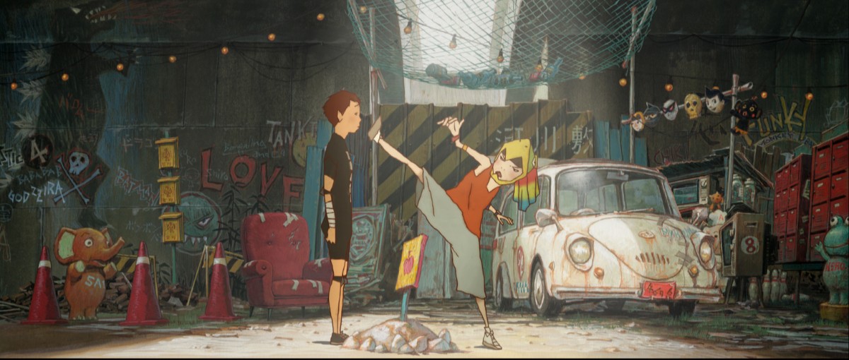 Two kids in a graffiti-covered junkyard; one girl kicks the other in a dynamic martial-arts pose next to a dusty vintage car and scattered props.